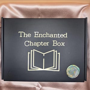 Puede incluir: Caja negra con letras doradas que dicen "The Enchanted Chapter Box". Un contorno dorado de un libro abierto y un sello de cera multicolor en la esquina inferior derecha.
