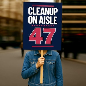 以下が含まれることがあります： 「CLEANUP ON AISLE」の文字と数字「47」が書かれた看板を持つ人。 看板は濃い青色で、白と赤の文字と星が描かれています。 その人はデニムジャケットを着ています。
