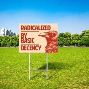 Op de afbeelding: Een rechthoekig bord met de tekst "RADICALIZED BY BASIC DECENCY" en een rode ravenillustratie. Het bord is gemonteerd op een wit metalen frame en staat in een grasveld onder een blauwe lucht.