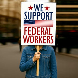 May include: A sign with the text "WE SUPPORT FEDERAL WORKERS" in red and blue, and an American flag graphic. The person holding the sign is wearing a blue denim jacket.