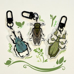 Puede incluir: Tres llaveros con temática de insectos con cierres negros. Los llaveros presentan un escarabajo azul, una cigarra verde y una mantis religiosa verde. Los insectos están representados con arte lineal detallado y están sobre un fondo blanco con acentos de hojas verdes.