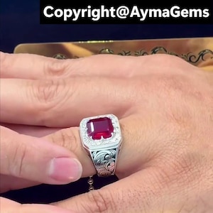 Peut inclure: Une bague en argent ornée d'une grosse pierre rouge carrée, entourée de petites pierres blanches. La bague présente un motif floral gravé sur le corps. Le texte "Copyright@AymaGems" est visible en haut.