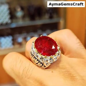 Peut inclure: Une bague ornée d'un grand rubis ovale. Le rubis est d'un rouge profond, serti dans une monture en métal argenté avec des détails complexes. La bague a un accent doré autour du rubis. Le texte dans l'image se lit : AymagemsCraft.