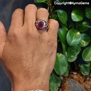 Peut inclure: Une bague en argent ornée d'un grand rubis ovale, entouré de petites pierres blanches. La bague présente un motif détaillé sur le corps. L'image contient également le texte "Copyright @AymaGems".