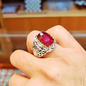 Peut inclure: Bague ornée d'un rubis de forme ovale. La bague présente un anneau argenté avec des détails de filigrane complexes et un sertissage doré. Le rubis, d'un rouge profond, est l'élément central de la bague.