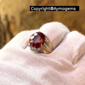 Peut inclure: Bague ornée d'un rubis rouge vif de forme ovale. La monture est en argent avec des détails dorés. Le texte "Copyright@Aymagems" est affiché dans un rectangle noir. La bague est présentée sur un fond blanc.