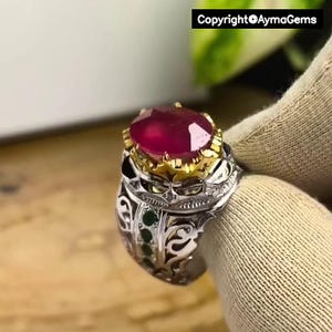 Peut inclure: Bague en argent ornée d'un grand rubis ovale. La bague présente un sertissage en or autour du rubis et des détails en filigrane argenté sur le corps. De petites pierres vertes sont serties sur les côtés. Copyright @AymaGems est visible.