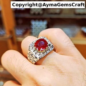 Peut inclure: Une bague en argent ornée d'une grosse pierre précieuse rouge foncé de forme ovale. La bague a une face carrée avec une bordure décorative et des détails en filigrane sur le corps. Le texte "Copyright@AymaGemsCraft" est en haut.