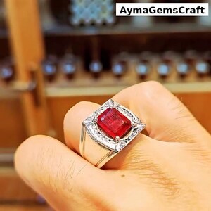 Peut inclure: Une bague en argent ornée d'une grande pierre rouge rectangulaire, entourée de petites pierres claires. La bague est portée sur une main, avec le texte "AymaGemsCraft" visible en arrière-plan.