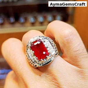 Peut inclure: Une bague en argent ornée d'une grande pierre rouge rectangulaire, entourée de petites pierres claires. La bague présente un design orné et détaillé. Le texte "AymaGemsCraft" est visible en haut à droite.