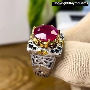 Peut inclure: Gros plan d'une bague ornée d'un grand rubis ovale. La bague a un sertissage en or autour du rubis et est ornée de pierres noires. Le corps de la bague est en argent avec des motifs de filigrane complexes. Le texte "Copyright@AymaGems" est visible.