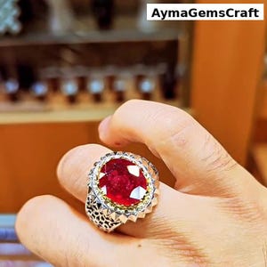 Peut inclure: Bague ornée d'un grand rubis ovale. La bague présente un anneau argenté avec des détails ajourés et un sertissage doré autour du rubis. Le texte "AymaGemsCraft" est visible en haut à droite.