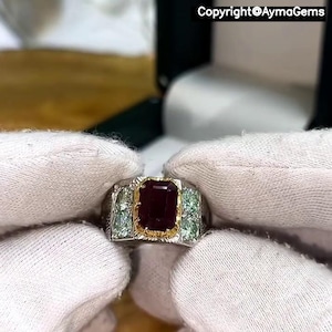 Peut inclure: Une bague en argent ornée d'une grande pierre rouge foncé rectangulaire, sertie d'or. Des pierres vertes claires plus petites encadrent la pierre centrale. Le texte "Copyright @AymaGems" est visible.