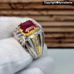 Peut inclure: Bague en argent et or ornée d'une grande pierre rouge foncé de forme rectangulaire. La bague est ornée de petites pierres bleues claires et est tenue dans une main gantée de blanc. Le texte "Copyright@AymaGems" est visible.