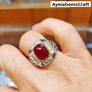 Peut inclure: Bague en argent ornée d'une grosse pierre rouge foncé de forme ovale. La pierre est entourée de petits brillants et d'un sertissage doré. La bague présente un motif ajouré ornemental sur le corps. Texte : AymagemsCraft.