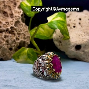 Peut inclure: Une bague en argent ornée d'une grosse pierre précieuse magenta ovale. La bague présente des détails complexes avec des accents dorés et de petites pierres de couleur turquoise. Le texte "Copyright@Aymagems" est visible en arrière-plan.