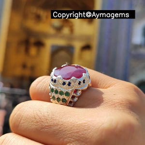 Peut inclure: Bague en argent ornée d'un grand rubis ovale. La bague est ornée de petites pierres bleues et vertes. Le texte "Copyright@Aymagems" est visible.