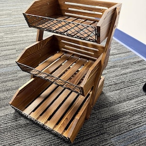 May include: A three-tiered wooden display rack with a rustic design. The rack features three open-top wooden trays with slatted bottoms and black wire mesh fronts. The wood has a warm, brown finish. The rack is designed to hold and display items.