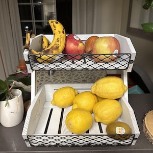 Op de afbeelding: Een witte houten fruitmand met twee niveaus en zwart gaas. De bovenste laag bevat bananen, rode appels en peren. De onderste laag bevat gele citroenen en een kiwi.
