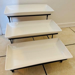 May include: A three-tiered serving stand with rectangular white ceramic platters. The platters are supported by a black metal frame. The stand is designed for displaying food items.