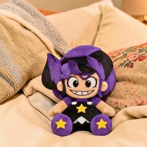Pode incluir: Uma boneca de pelúcia com um design de desenho animado. A boneca tem um penteado roxo e preto, um rosto sorridente e está vestida com uma roupa preta com detalhes de estrelas amarelas. Os pés da boneca são roxos com detalhes de estrelas amarelas. A boneca está sentada em uma cama.