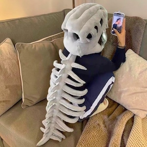 May include: A person wearing a grey dinosaur skull headpiece and a matching rib cage costume, taking a selfie with a smartphone. The costume is made of soft, plush material. The person is sitting on a brown sofa with cushions.