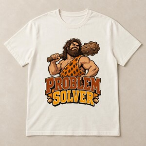 May include: A cream-coloured short-sleeve t-shirt featuring a graphic of a muscular caveperson with a beard, wearing a leopard-print tunic and holding a wooden club. Below the figure, the text reads "PROBLEM SOLVER" in bold, orange-outlined letters.