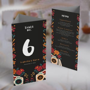 May include: Black table number card with a large white number 6. The card features floral and sugar skull designs in red, orange, and teal. The text "Table No. 6" and names "Isabella & Maria" are visible. A menu card is also shown.