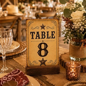 May include: A rectangular table number card with a vintage parchment-style finish, featuring the text "TABLE 8" in black lettering, flanked by two star icons, and held upright by a dark brown wooden base with decorative scrollwork borders.