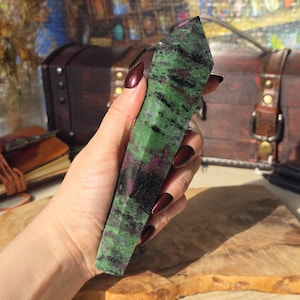 Può includere: Una punta di cristallo verde, nero e rosa, nota anche come pietra preziosa, a forma di una lunga piramide sottile a sei lati. Il cristallo è tenuto in una mano.