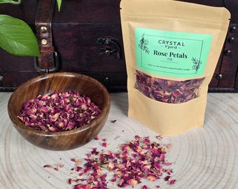 May include: A brown paper bag with a green label that reads "CRYSTAL - Fjord - Rose Petals 25g". The bag contains dried rose petals. A wooden bowl filled with dried rose petals sits on a light wood surface.