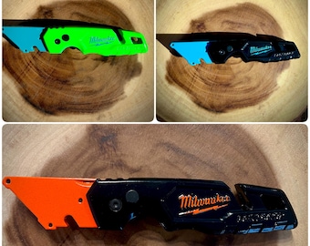 May include: Three Milwaukee utility knives with different colored handles. The first knife has a green handle, the second knife has a black handle, and the third knife has an orange handle. All three knives have a blue blade and the Milwaukee logo on the side.