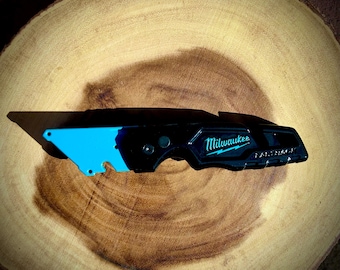 May include: A black and blue utility knife with a retractable blade. The knife has a black handle with the word "Milwaukee" printed on it in white letters. The blade is partially extended and is blue.