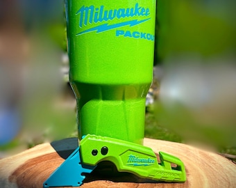 May include: A bright green tumbler and utility knife set. The tumbler features the Milwaukee logo in blue. The utility knife is also green with a blue blade and the Milwaukee logo. The items are on a wooden surface.