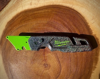 May include: A utility knife with a neon green blade and a textured, glittery black and gray handle. The Milwaukee logo is visible in green. The knife is resting on a wooden surface.