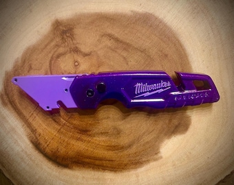 May include: A purple utility knife with the Milwaukee logo in white. The knife has a two-tone design, with a lighter purple blade section and a darker purple handle. The knife is lying on a wooden surface.
