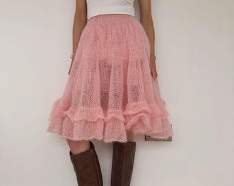 May include: A light pink, ruffled skirt paired with a white tank top and brown knee-high boots. The skirt has a layered design, and the boots have a buckle detail. The outfit is set against a plain white wall.