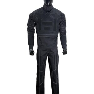 May include: A black tactical-style outfit consisting of a long-sleeved top with a high neck, padded shoulder panels, and rectangular hook-and-loop patches, paired with matching trousers. The garment features prominent white contrast stitching.