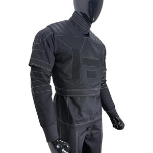 May include: A black long-sleeved shirt and trousers set featuring a high collar, quilted shoulder panels, and white contrast stitching. The garment includes rectangular hook-and-loop fastener patches on the chest and legs, with ribbed cuffs.