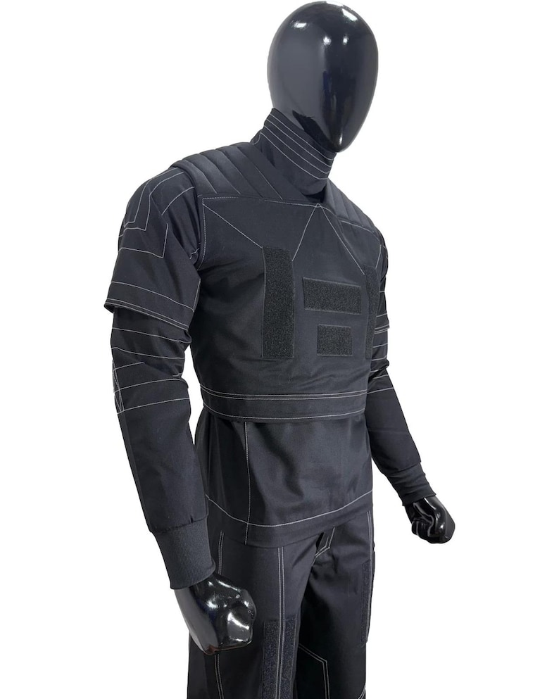 May include: A black long-sleeved shirt and trousers set featuring a high collar, quilted shoulder panels, and white contrast stitching. The garment includes rectangular hook-and-loop fastener patches on the chest and legs, with ribbed cuffs.