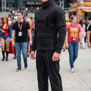 May include: A black long-sleeved mock neck shirt and matching black trousers featuring white contrast stitching in geometric patterns. The shirt has a high collar and a structured design, while the trousers are straight-legged and black.