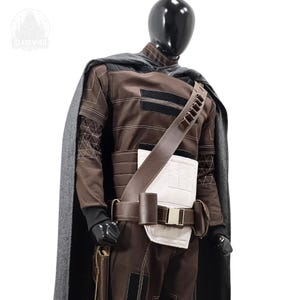 May include: A brown and black costume set featuring a long grey cape, a brown jacket with white paneling, and a brown utility belt with pouches. The outfit includes a diagonal shoulder strap with ammunition loops and black accents throughout.