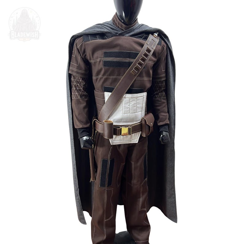 May include: A brown costume set featuring a long gray cape, a brown long-sleeved top with black accents, brown trousers, a white chest panel, and a brown utility belt with a gold buckle and attached pouches, worn by a black mannequin.