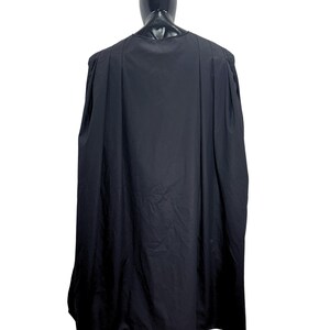 May include: A solid black cape featuring a rounded neckline and vertical pleats that extend from the shoulders down the length of the garment. The fabric drapes loosely, creating a full silhouette that covers the back and shoulders of the mannequin.