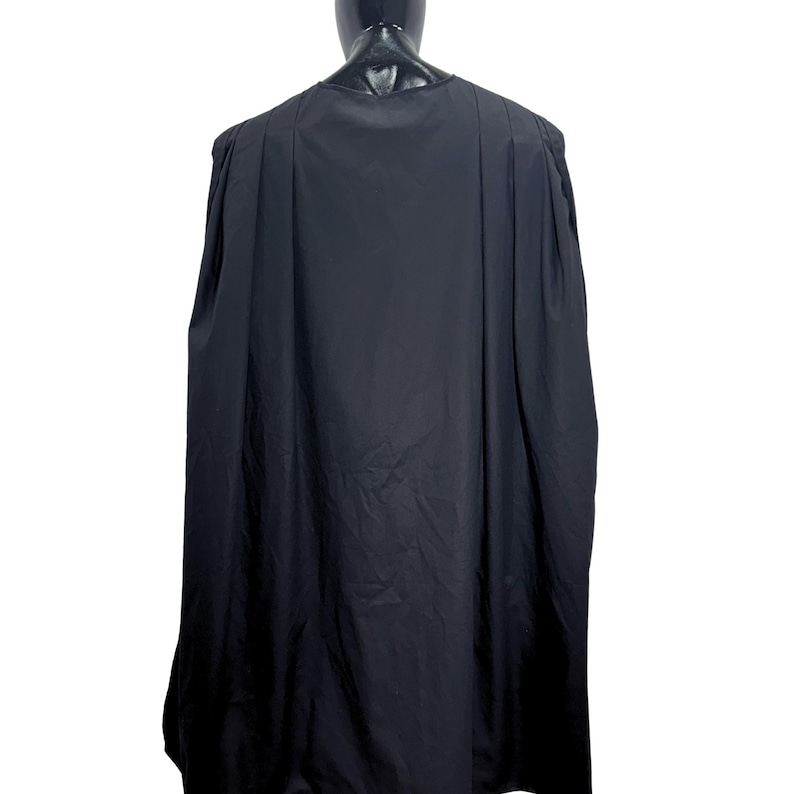 May include: A solid black cape featuring a rounded neckline and vertical pleats that extend from the shoulders down the length of the garment. The fabric drapes loosely, creating a full silhouette that covers the back and shoulders of the mannequin.