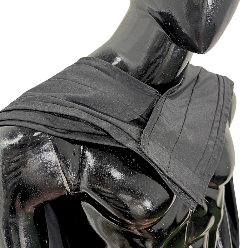 May include: A black fabric shoulder wrap featuring a structured, pleated panel on one side and a soft, draped section on the other. The garment is displayed on a glossy black mannequin, showcasing the contrast between the smooth and textured fabric.