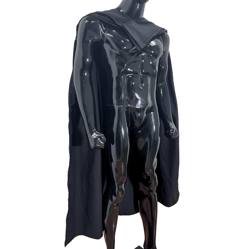 May include: A solid black cape draped over a glossy black mannequin. The cape features a structured collar section that rests across the shoulders and extends down the back, providing full coverage with a smooth, matte fabric finish throughout.