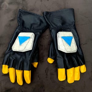 May include: A pair of black leather gloves with yellow fingertips. Each glove features a white and silver patch with a blue triangle. The gloves are presented on a brown surface.