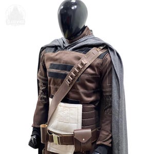 May include: A brown long-sleeved tunic with white rectangular panels, a grey cape, and a brown utility belt featuring a holster and pouch. A diagonal strap with small cylindrical loops crosses the chest. The outfit is displayed on a black mannequin.