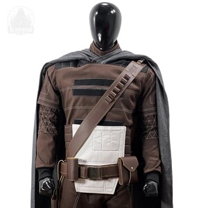 May include: A brown long-sleeved tunic with a white chest panel, a grey cape, and a brown utility belt featuring a diagonal shoulder strap with ammunition loops. The outfit is displayed on a black mannequin with a logo reading BLADEWISH.